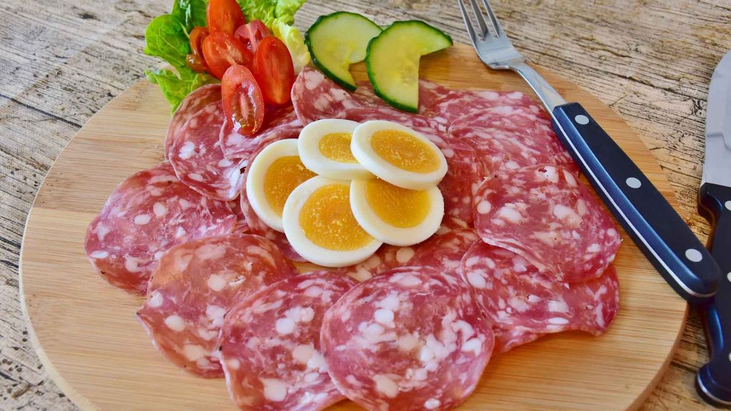 Sliced sausage, eggs, and some vegetables are arranged in a circle atop a wooden plate