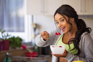 A smiling woman, holding a bowl filled with berries and a spoon filled with 2 spoons, ready to eat.